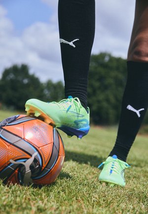 Joueur de football debout sur l'herbe avec un pied en crampons bleu-vert reposant sur un ballon de football orange et noir en extérieur.