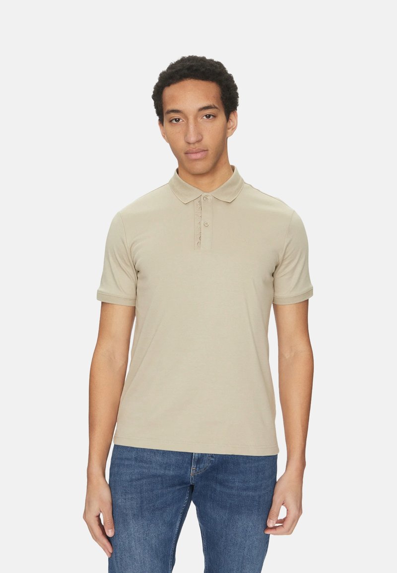 Polo beige realizzato in morbido tessuto, con colletto classico, patta con tre bottoni e maniche corte, abbinato a jeans blu.