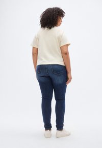 Κρεμ κοντομάνικη μπλούζα σε συνδυασμό με σκούρο μπλε στενά τζιν και λευκά αθλητικά παπούτσια, από πίσω. Απλός σχεδιασμός, casual εφαρμογή.