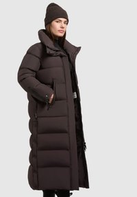 Cappotto imbottito lungo di colore marrone scuro, con colletto alto, tasche laterali con zip e chiusura a bottoni. Texture liscia con sezioni trapuntate.