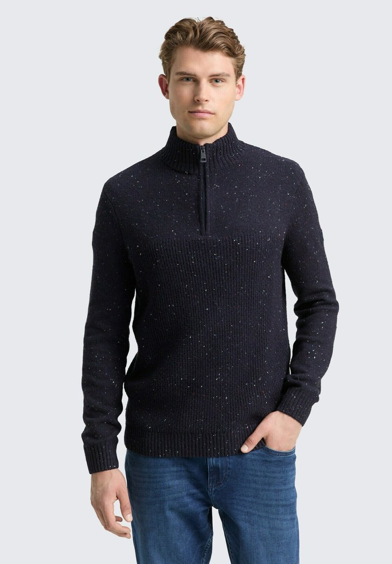 Mann trägt einen dunkel gesprenkelten Strickpullover mit halbem Reißverschluss und blaue Jeans, steht mit einer Hand in der Tasche vor einem hellen Hintergrund.