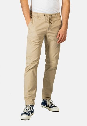 Beige Baumwollmischungshosen mit elastischem Bund und Kordelzug, mit zwei seitlichen Taschen und schlankem Schnitt. Kombiniert mit blauen Sneakers.