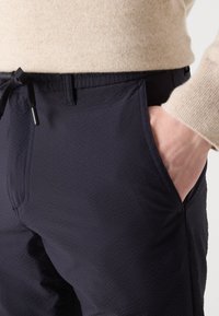 Gros plan sur un pantalon texturé bleu marine avec un cordon noir, des poches avant et une manche de pull beige reposant sur la hanche.