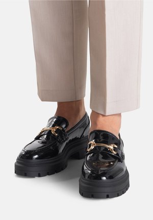 Mocasines de charol negro con suelas gruesas de tipo lug y detalles metálicos dorados en forma de freno de caballo, usados con pantalones beige que terminan por encima de los tobillos.