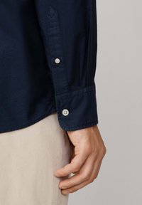 La mano de un hombre descansando a un lado, vistiendo una camisa de manga larga azul marino con puños abotonados y pantalones beiges, contra un fondo neutral.