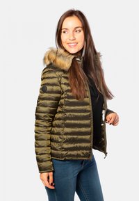 Doudoune vert olive avec une capuche en fausse fourrure, design matelassé et fermeture éclair. Assortie avec un haut noir et un jean foncé.