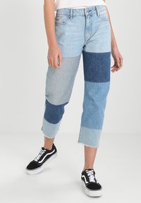 Ljusblå jeans med lapptäcktsdesign som har olika nyanser av denim. Kort längd med fransiga fållar, parat med svarta sneakers.