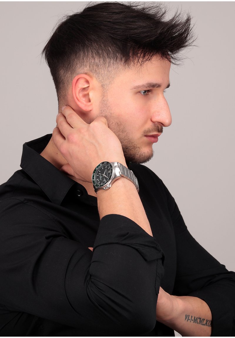 Orologio da polso in metallo argentato con quadrante nero e indicatori luminosi, indossato su una mano che riposa contro il collo. Camicia nera con maniche arrotolate.