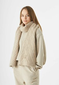 Jonge vrouw met lang haar draagt een beige brei-sjaal, een bijpassende oversized trui en broek, met de handen in de zakken tegen een witte achtergrond.