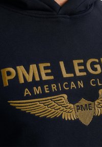 Granatowy hoodie z haftowanym złotym napisem "PME LEGEND AMERICAN CLUB", ozdobiony skrzydlatym wzorem i akcentami w kształcie tarczy.