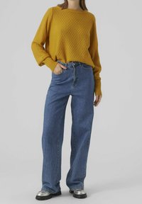 Gelber, strukturierter Strickpullover mit langen Ärmeln, kombiniert mit hoch taillierten, blauen Denim-Jeans mit geradem Bein und Schuhen mit silbernen Akzenten.