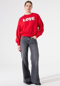 Sudadera roja con texto blanco "LOVE", de corte relax; combinada con jeans acampanados gris oscuro y botas negras sobre un fondo sencillo.