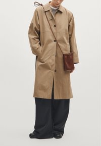 Beżowy, oversize płaszcz z kołnierzem, z przodu na guziki i bocznymi kieszeniami. Zestawiony z czarnymi spodniami typu wide-leg oraz małą brązową skórzaną torbą na ramię.