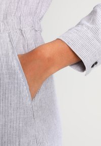 Main droite glissée dans une poche latérale verticale d'un vêtement rayé gris clair et blanc avec manches longues retroussées et taille froncée.