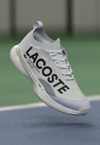 Lacoste idrettssko i lys grå med en glatt overdel, forsterket tå, dristig svart logo og polstret såle, designet for sportsytelse.