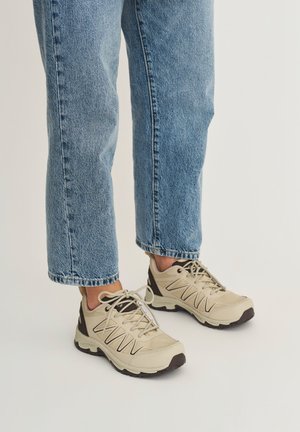 Persoon die lichtbeige sportschoenen met zwarte accenten en lichtblauwe jeans met rechte pijpen draagt, staand op een effen achtergrond.