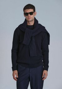 Navy-blau gestrickter Pullover mit Zopfmuster, kombiniert mit einem passenden Schal, der lässig über die Schultern gelegt ist, und dazu maßgeschneiderte dunkle Hose.