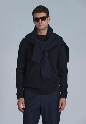 Marineblauwe gebreide sweater met kabelpatroon, gecombineerd met een bijpassende sjaal die over de schouders hangt, ter aanvulling van op maat gemaakte donkere broeken.
