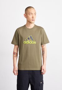 T-shirt en coton vert olive avec un grand logo multicolore d'Adidas sur le devant. Manches courtes et col rond. Coupe décontractée.