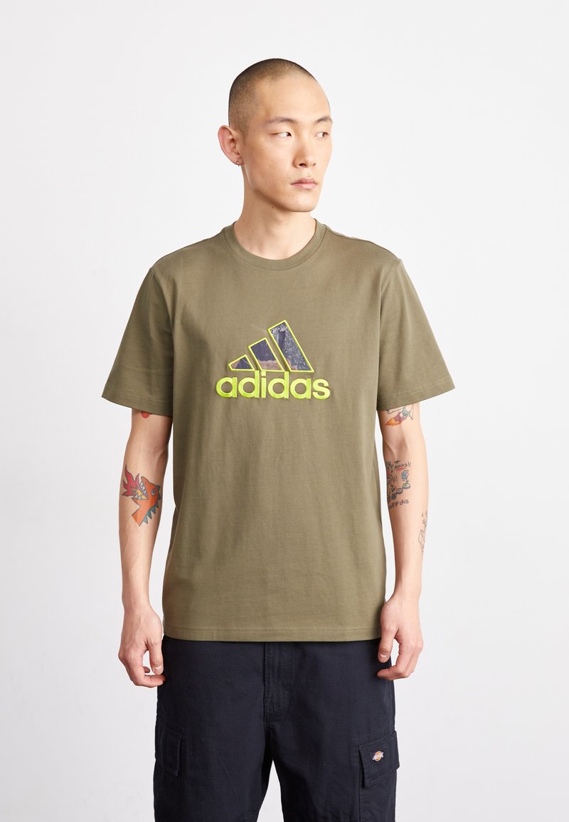T-shirt en coton vert olive avec un grand logo multicolore d'Adidas sur le devant. Manches courtes et col rond. Coupe décontractée.