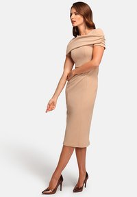 Beige bodycon dress with a draped neckline and short sleeves. Smooth texture, knee-length, paired with glossy brown stilettos.