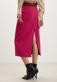 Gonna a matita fucsia con spacco posteriore, dotata di una cintura nera con dettagli borchiati. Tessuto morbido, design alla ginocchio, abbinata a stivali neri con tacco.
