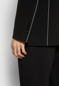 Veste de costume noire avec surpiqûres blanches contrastantes, trois boutons foncés sur le poignet, portée avec une bague transparente à la main.