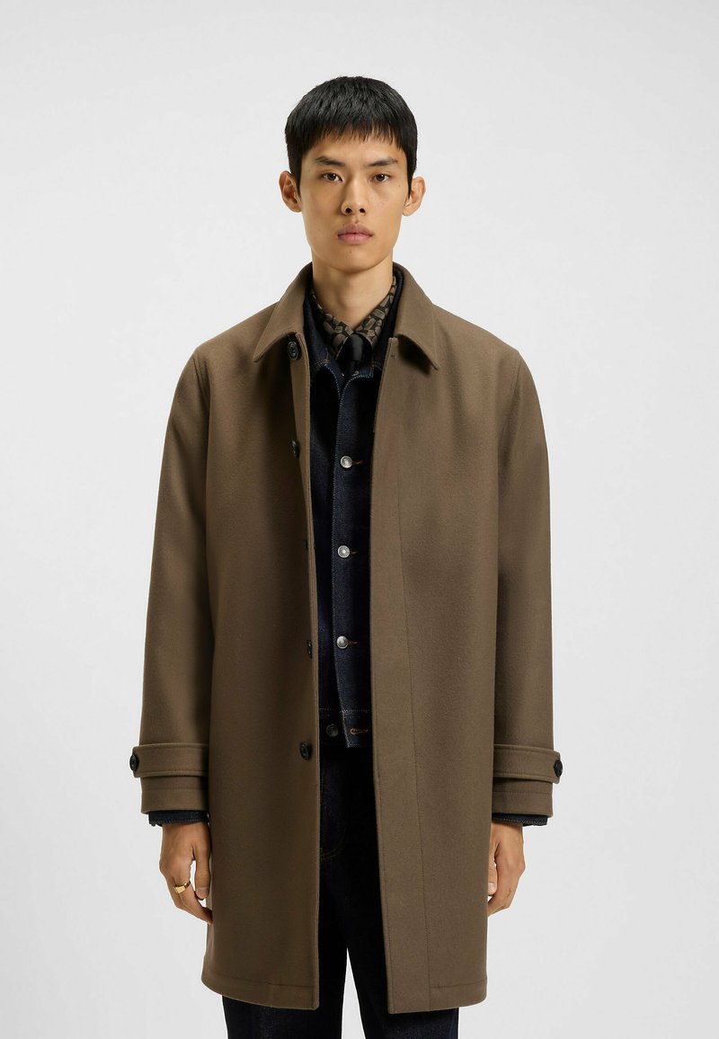 Brun uldblanding overfrakke med krave, knaplukning og to sidelommer. Bæres over en mørk denimskjorte.