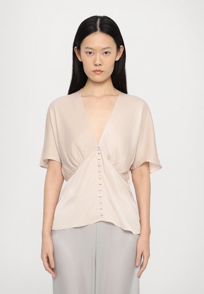 Beige silk blouse with short sleeves, deep V-neck, gathered waist, and a front button closure. Smooth texture and flowing design.