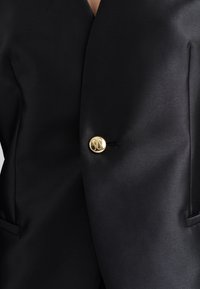 Blazer noir avec un tissu texturé, un décolleté en V profond, et un détail de bouton en or proéminent au centre.