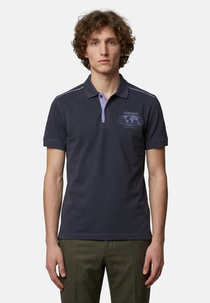 Junger Mann mit lockigem Haar, der ein dunkelblaues Polohemd mit Weltkartengrafik und olivgrüne Hosen vor neutralem Hintergrund trägt.