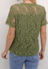 Haut en dentelle vert olive avec encolure festonnée et manches courtes, présentant un motif floral et un design superposé, porté avec un jean bleu.