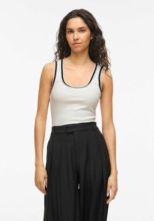 Kobieta o kręconych włosach, ubrana w biały bezrękawnik z czarnym wykończeniem i czarne plisowane spodnie z wysokim stanem, stojąca na białym tle.