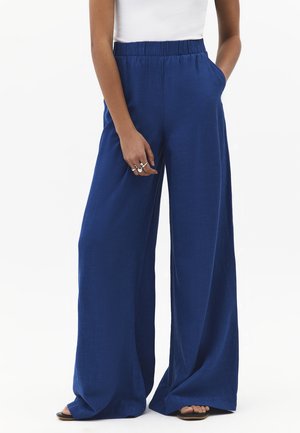 Femme portant un pantalon ample à jambes larges bleu marine avec taille élastique, un haut blanc sans manches et des bagues argentées, debout avec une main dans la poche.