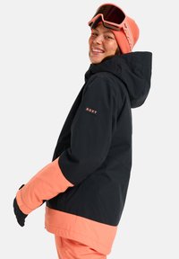 Jachetă de schi neagră și corai, cu glugă, având logo-ul "ROXY" pe mânecă, mobilitate amplă pentru brațe și manșete texturate.