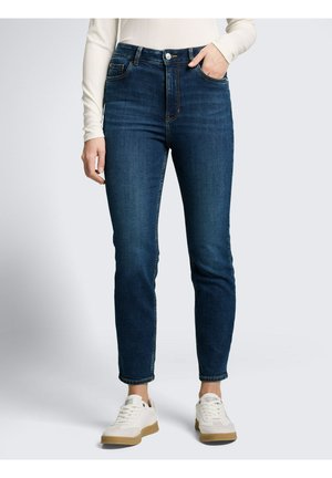 Vrouw draagt donkerblauwe slim-fit jeans, witte top met lange mouwen en witte sneakers met rubberen zolen, staat met één hand in de zak.