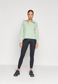 Chaqueta verde claro con cremallera y detalles reflectantes, combinada con leggings negros y zapatillas deportivas negras. Material suave y ligero.