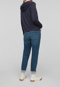 Marinblå hoodie med detalj på bakseam, tillsammans med jeans med uppvikta ärmar och vita högskor, placerad mot en enfärgad bakgrund.