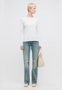 Γυναίκα με κοντά ξανθά μαλλιά που φοράει λευκό πουκάμισο με μακριά μανίκια, μπλε τζιν, μπεζ παπούτσια, κρατώντας μπεζ τσάντα, κοιτάζοντας προς το πλάι.