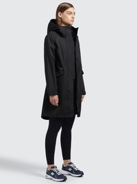 Musta vedenpitävä takki hupulla, etuvetoketjulla ja napilla. Yhdistetty mustiin leggings-housuihin ja sinisiin urheilukenkään. Yksinkertainen muotoilu, jossa on taskut.