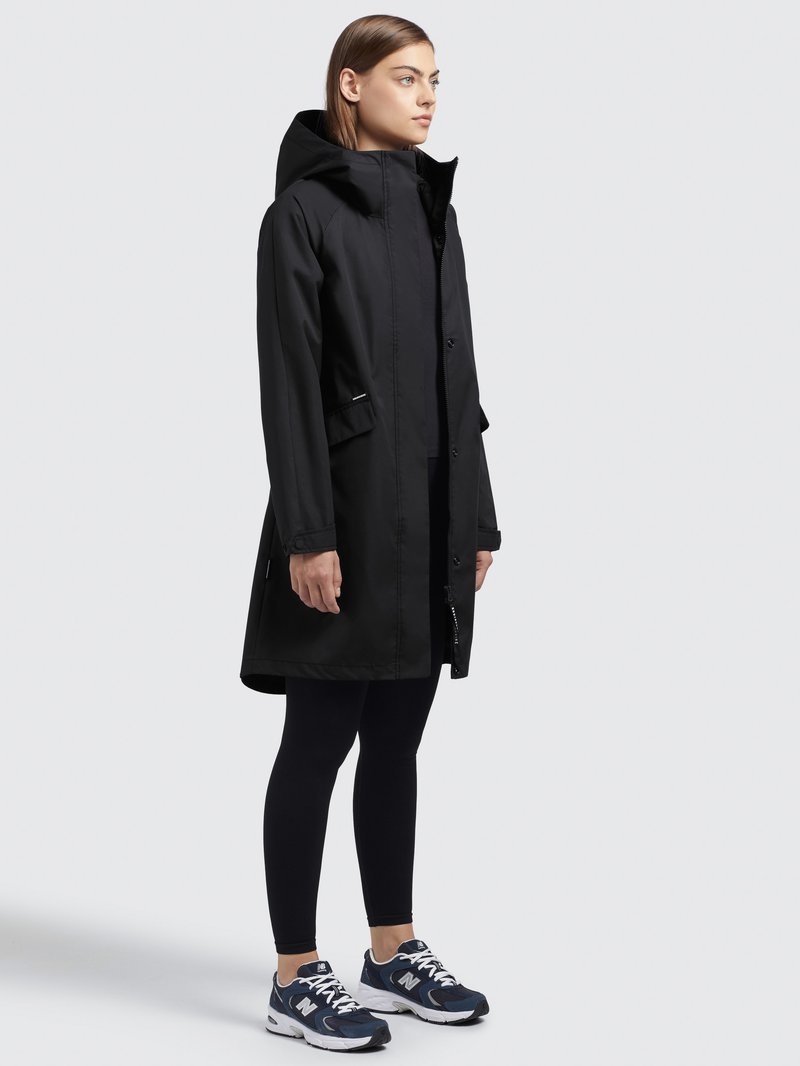 Veste imperméable noire avec capuche, zip frontal et boutons-pression. Associée à des leggings noirs et des chaussures de sport bleues. Design simple avec poches.