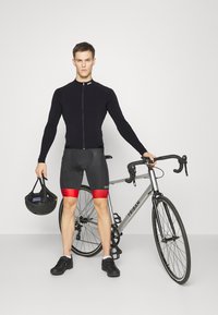 Cycliste en maillot à manches longues noir et short gris/rouge, tenant un casque noir, à côté d'un vélo argenté avec des guidons tombants.