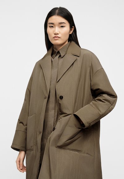 Fiatal nő hosszú fekete hajjal, túlméretezett barna kabátot és hozzáillő inget visel, egyszínű fehér háttér előtt.