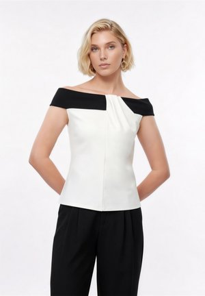 Mujer con cabello rubio corto, vestida con un top negro y blanco sin hombros y pantalones negros, de pie con las manos detrás de la espalda sobre un fondo blanco.