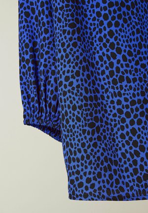 Tessuto blu con un motivo a macchie nere, con polsini delle maniche arricciati. La trama appare liscia e leggera.