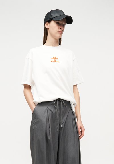 Γυναίκα που φοράει λευκό μπλουζάκι με πορτοκαλί κείμενο "ΕΙΝΑΙ ΚΑΛΥΤΕΡΑ ΣΤΟ STIEGLITZ", γκρι πλισέ παντελόνι με κορδόνι και μαύρο δερμάτινο καπέλο бейζбол.