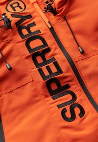 Pomarańczowa kurtka z czarnym, wyhaftowanym napisem "SUPERDRY®", czarnym zamkiem błyskawicznym oraz czarnymi sznureczkami ze plastikowymi regulatorami.