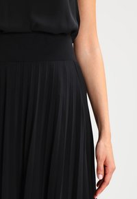 Femme portant une jupe plissée noire et un haut noir, avec le bras droit détendu à ses côtés, contre un fond clair.