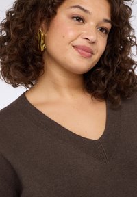 Maglione scollo a V marrone scuro realizzato in tessuto morbido. Presenta una maglia a trama e una leggera costina lungo il collo. Accenti di design minimalisti.