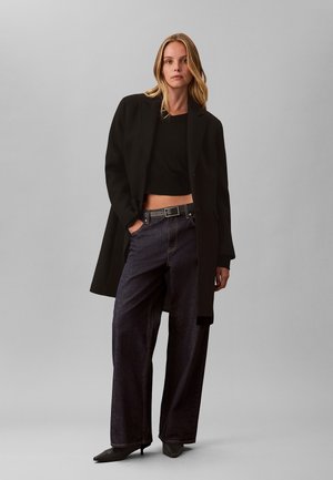 Femme aux longs cheveux blonds portant un long manteau noir, un crop top noir, un jean large foncé, une ceinture noire et des talons pointus noirs, debout devant un fond gris.
